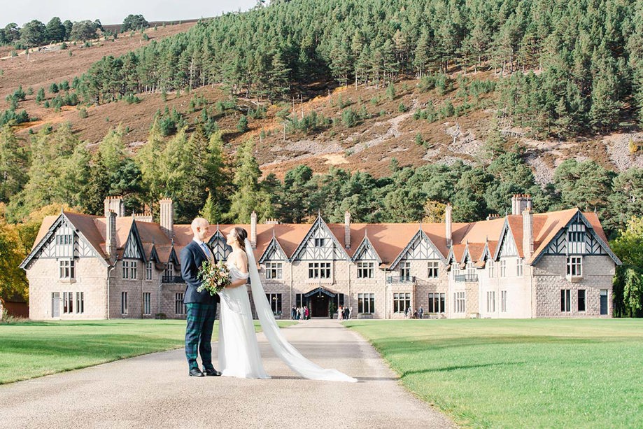 bride and groom standing in front of large country house with Scottish hills surrounding them